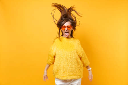 Funny girl in fur sweater, heart orange glasses showing tongue, fooling around in studio jump with fluttering hair isolated on yellow background. People sincere emotions, lifestyle. Advertising areaの写真素材
