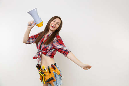 Young excited handyman woman in plaid shirt, denim shorts, kit tools belt full of variety instruments hold megaphone, screaming isolated on white background. Female in male work. Renovation conceptの写真素材