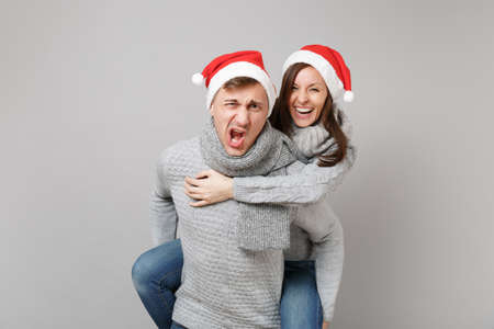 Merry fun couple girl guy in red Santa Christmas hat gray sweaters scarves isolated on grey wall background, studio portrait. Happy New Year 2019 celebration holiday party concept. Mock up copy spaceの写真素材