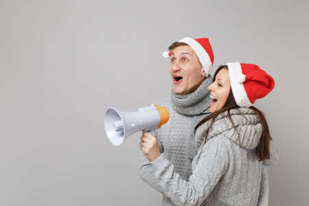 Merry fun couple girl guy in red Santa Christmas hat gray sweaters scarves isolated on grey wall background, studio portrait. Happy New Year 2019 celebration holiday party concept. Mock up copy spaceの写真素材