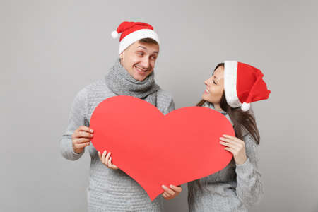 Merry fun couple girl guy in red Santa Christmas hat gray sweaters scarves isolated on grey wall background, studio portrait. Happy New Year 2019 celebration holiday party concept. Mock up copy spaceの写真素材