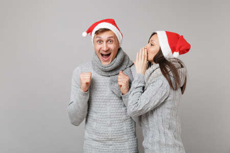 Merry fun couple girl guy in red Santa Christmas hat gray sweaters scarves isolated on grey wall background, studio portrait. Happy New Year 2019 celebration holiday party concept. Mock up copy spaceの写真素材