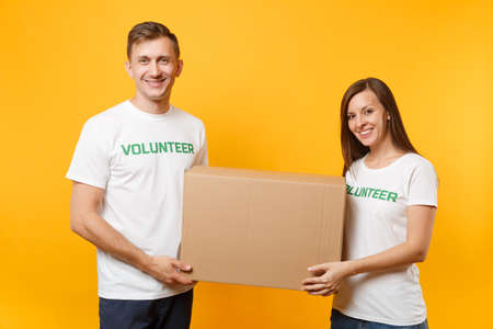 Colleagues couple in white t-shirt with written inscription green volunteer hold cardboard box donations isolated on yellow background. Voluntary free assistance help, charity grace teamwork conceptの写真素材