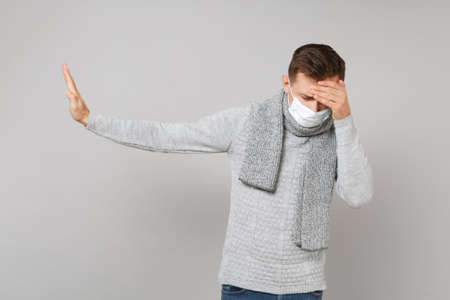 Young man in sterile face mask with lowered head putting hands on forehead, showing stop gesture with palm aside isolated on grey background. Healthy, ill sick disease treatment, cold season conceptの写真素材