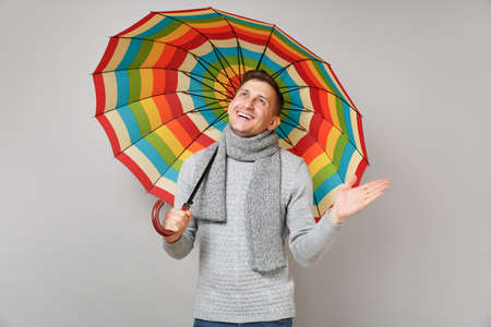 Joyful young man in gray sweater, scarf looking up, holding colorful umbrella, spreading hand isolated on grey wall background. Healthy fashion lifestyle, people sincere emotions, cold season conceptの写真素材
