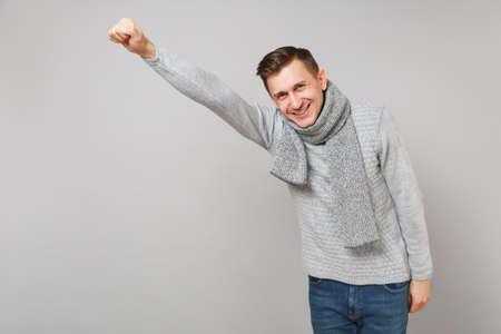 Cheerful young man in gray sweater, scarf with outstretched hand like Superman isolated on grey background. Healthy fashion lifestyle, people sincere emotions, cold season concept. Mock up copy spaceの写真素材