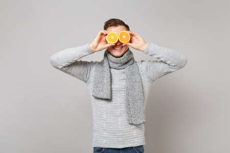 Smiling young man in gray sweater, scarf covering eyes with oranges isolated on grey background in studio. Healthy fashion lifestyle, people sincere emotions, cold season concept. Mock up copy spaceの写真素材