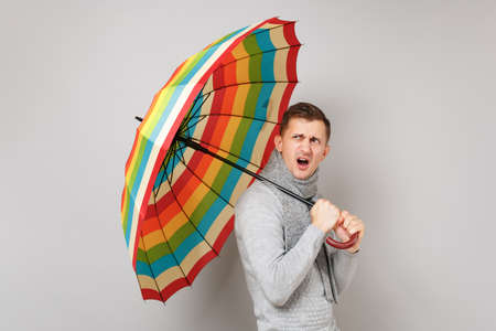 Irritated young man in gray sweater, scarf holding colorful umbrella isolated on grey background in studio. Healthy fashion lifestyle, people sincere emotions, cold season concept. Mock up copy spaceの写真素材