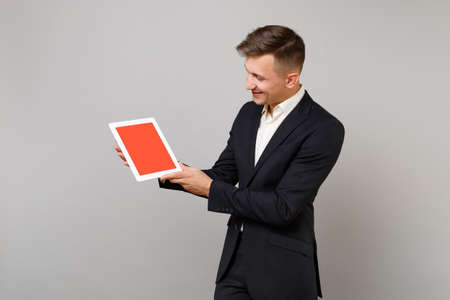 Young business man in classic black suit holding and looking on tablet pc computer with blank empty screen isolated on grey background. Achievement career wealth business concept. Mock up copy spaceの写真素材