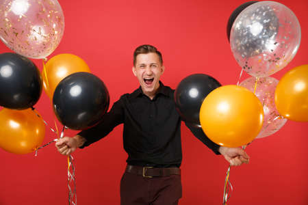 Happy young man in black classic shirt holding air balloons, celebrating isolated on bright red background. Valentine's, International Women's Day Happy New Year birthday mockup holiday party conceptの写真素材