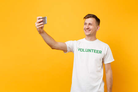 Portrait of man in white t-shirt written inscription green title volunteer taking selfie shot on mobile phone isolated on yellow background. Voluntary free assistance help charity grace work concept