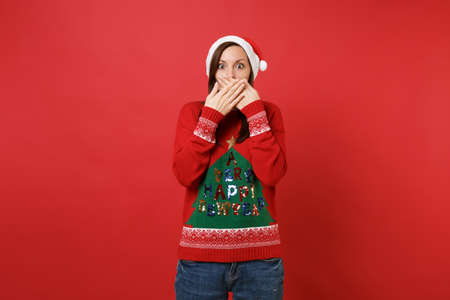 Shocked young Santa girl in knitted sweater, Christmas hat covering mouth with hands isolated on bright red wall background. Happy New Year 2019 celebration holiday party concept. Mock up copy spaceの写真素材