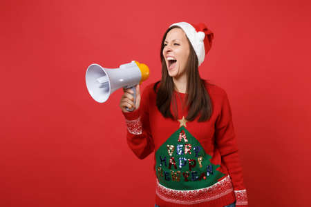 Irritated young Santa girl in knitted sweater, Christmas hat screaming on megaphone isolated on bright red wall background. Happy New Year 2019 celebration holiday party concept. Mock up copy spaceの写真素材