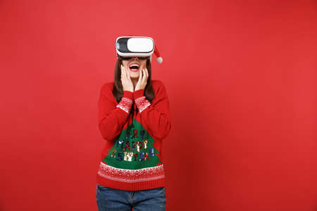 Amazed young Santa girl in knitted sweater, Christmas hat looking in headset, put hands on face...