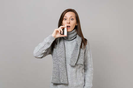 Shocked young woman in gray sweater, scarf holding using nasal drops isolated on grey wall background in studio. Healthy lifestyle, ill sick disease treatment, cold season concept. Mock up copy spaceの写真素材