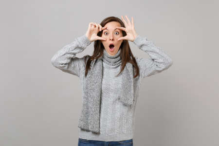 Shocked young woman in gray sweater, scarf keeping mouth wide open, stretching eyelids isolated on grey background. Healthy fashion lifestyle, people emotions, cold season concept. Mock up copy spaceの写真素材