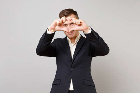 Portrait of joyful young business man in classic black suit, shirt showing shape heart with hands isolated on grey background in studio. Achievement career wealth business concept. Mock up copy spaceの写真素材