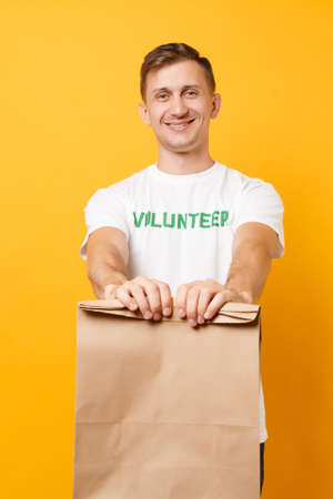 Portrait of man in white t-shirt written inscription green title volunteer hold blank craft paper bag for takeaway isolated on yellow background. Voluntary free assistance help charity grace conceptの写真素材