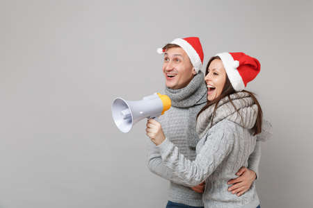 Merry fun couple girl guy in red Santa Christmas hat gray sweaters scarves isolated on grey wall...