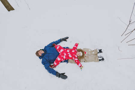 Happy family man, little girl in colorful warm clothes spreading hands, making snow angels while lying in park or forest outdoors. Winter fun, leisure on holidays. Love relationship lifestyle conceptの写真素材