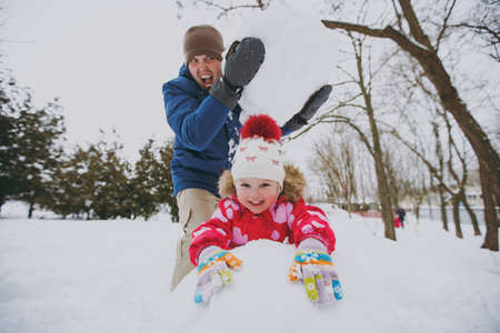 Funny family screaming dad in warm clothes holding big snowball, little girl playing with snow in park or forest outdoors. Winter fun, leisure on holidays. Love relationship family lifestyle conceptの写真素材