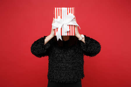 Woman in fur sweater covering face with red striped present box with gift ribbon isolated on red...