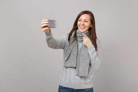 Smiling girl in gray sweater scarf pointing index finger doing selfie shot on mobile phone making video call isolated on grey background. Healthy fashion lifestyle people emotions cold season conceptの写真素材