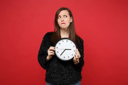 Puzzled young woman in black fur sweater looking up, biting lips, holding round clock isolated on bright red wall background in studio. People sincere emotions, lifestyle concept. Time is running outの写真素材