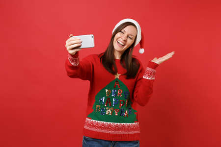 Joyful Santa girl in Christmas hat doing taking selfie shot on mobile phone, pointing hand aside isolated on red background. Happy New Year 2019 celebration holiday party concept. Mock up copy spaceの写真素材