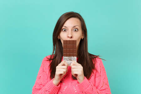 Shocked young woman in knitted pink sweater hold in hands, covering mouth with chocolate bar...