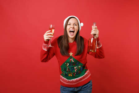 Overjoyed young Santa girl in Christmas hat screaming, holding glass bottle of champagne isolated on bright red background. Happy New Year 2019 celebration holiday party concept. Mock up copy spaceの写真素材