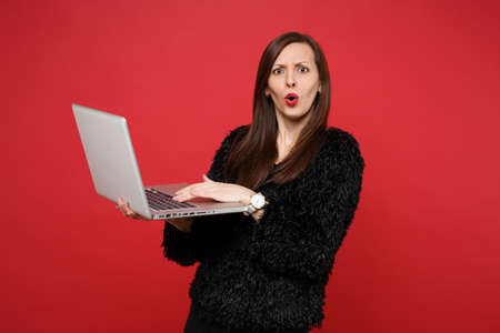 Shocked perplexed young woman in black fur sweater hold, working on laptop pc computer isolated on...