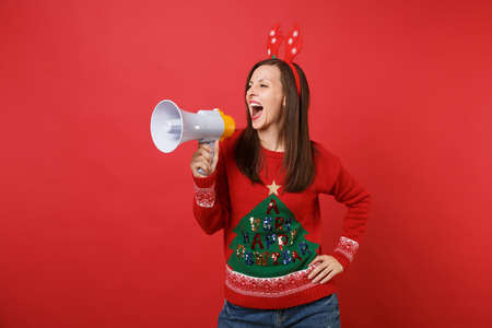 Emotional young Santa girl in fun decorative deer horns on head screaming on megaphone isolated on bright red background. Happy New Year 2019 celebration holiday party concept. Mock up copy spaceの写真素材