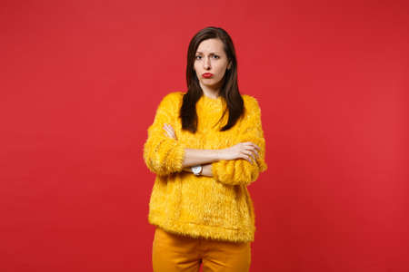 Portrait of upset sad offended young woman in yellow fur sweater holding hands folded isolated on bright red wall background in studio. People sincere emotions, lifestyle concept. Mock up copy spaceの写真素材