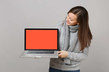 Curious young woman in gray sweater, scarf hold looking on laptop pc computer with blank empty screen isolated on grey background. Healthy lifestyle, online treatment consulting, cold season conceptの写真素材