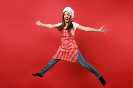 Housewife female chef cook or baker in striped apron white t-shirt, toque chefs hat isolated on red wall background. Full length portrait housekeeper woman jumping high up. Mock up copy space conceptの写真素材