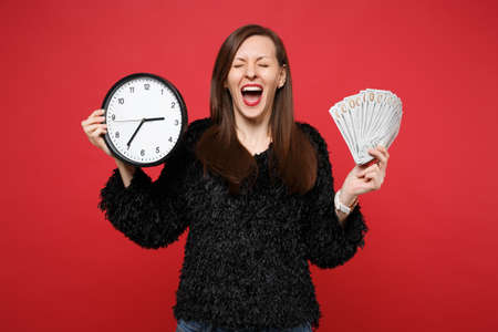 Happy woman in black fur sweater with closed eyes, opened mouth holding round clock, fan of money in...