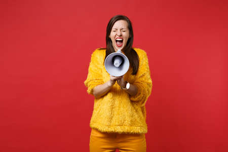 Crazy young woman in yellow fur sweater keeping eyes closed, screaming on megaphone isolated on bright red wall background in studio. People sincere emotions, lifestyle concept. Mock up copy spaceの写真素材