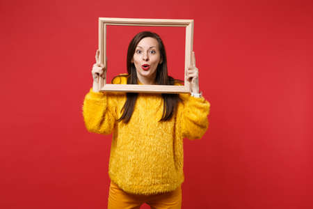 Portrait of amazed attractive young woman in yellow fur sweater holding picture frame isolated on bright red wall background in studio. People sincere emotions, lifestyle concept. Mock up copy spaceの写真素材