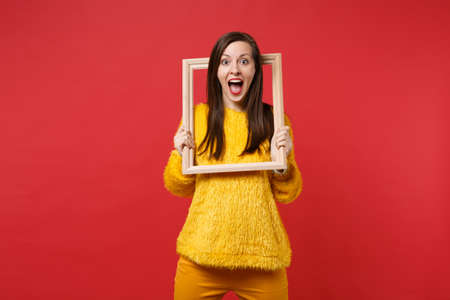 Portrait of surprised young woman in yellow fur sweater keeping mouth wide open holding picture...