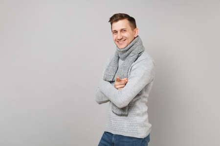 Smiling young man in gray sweater, scarf holding hands folded pointing index finger on camera isolated on grey wall background. Healthy fashion lifestyle, people sincere emotions, cold season conceptの写真素材