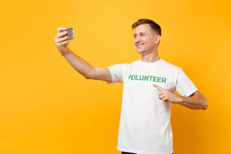 Portrait of man in white t-shirt written inscription green title volunteer taking selfie shot on mobile phone isolated on yellow background. Voluntary free assistance help charity grace work conceptの写真素材