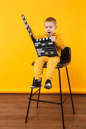 Little kid boy 3-4 years old in yellow clothes isolated on orange wall background, children studio portrait. People childhood lifestyle concept. Hand hold film making clapperboard. Mock up copy spaceの写真素材