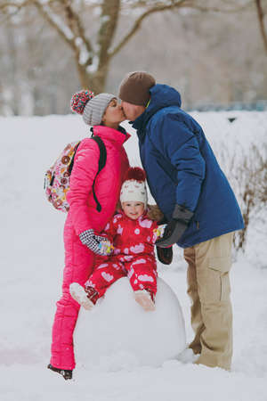 Young family woman, man in warm clothes kissing, little girl sitting on snowball in snowy park or...