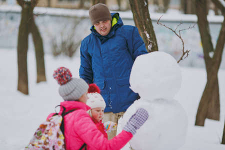 Young family man woman, little girl in winter warm clothes playing, making snowman in snowy park or...
