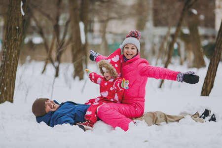 Laughing family woman, man little girl in warm clothes lying, sitting on snow, spreading hands in park or forest outdoors. Winter fun, leisure on holidays. Love relationship people lifestyle conceptの写真素材