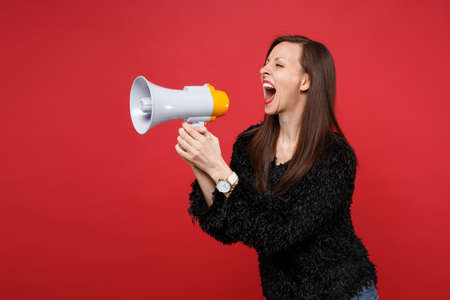 Portrait of crazy young girl in black fur sweater looking aside screaming on megaphone isolated on...