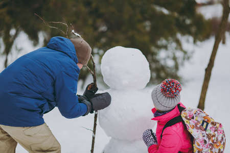 Back rear view of couple woman, man in winter warm clothes playing, making snowman in snowy park or forest outdoors. Winter fun, leisure on holidays. Love relationship family people lifestyle conceptの写真素材