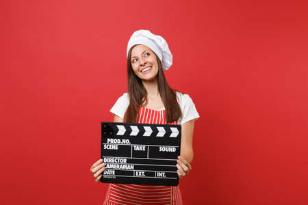 Housewife female chef cook or baker in striped apron, white t-shirt, toque chefs hat isolated on red wall background. Woman holding classic black film making clapperboard. Mock up copy space conceptの写真素材