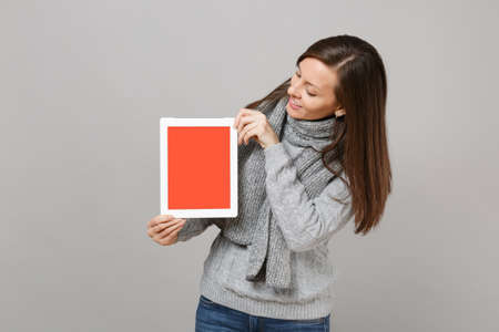 Young woman in sweater, scarf looking on tablet pc computer with blank empty screen isolated on grey background. Healthy lifestyle, online treatment consulting cold season concept. Mock up copy spaceの写真素材